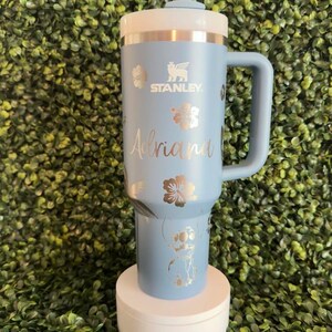 May include: A light blue Stanley tumbler with a white lid and handle. The tumbler features silver floral designs, the name "Adriana", and a cartoon character. It includes a white straw and is displayed on a white stand.