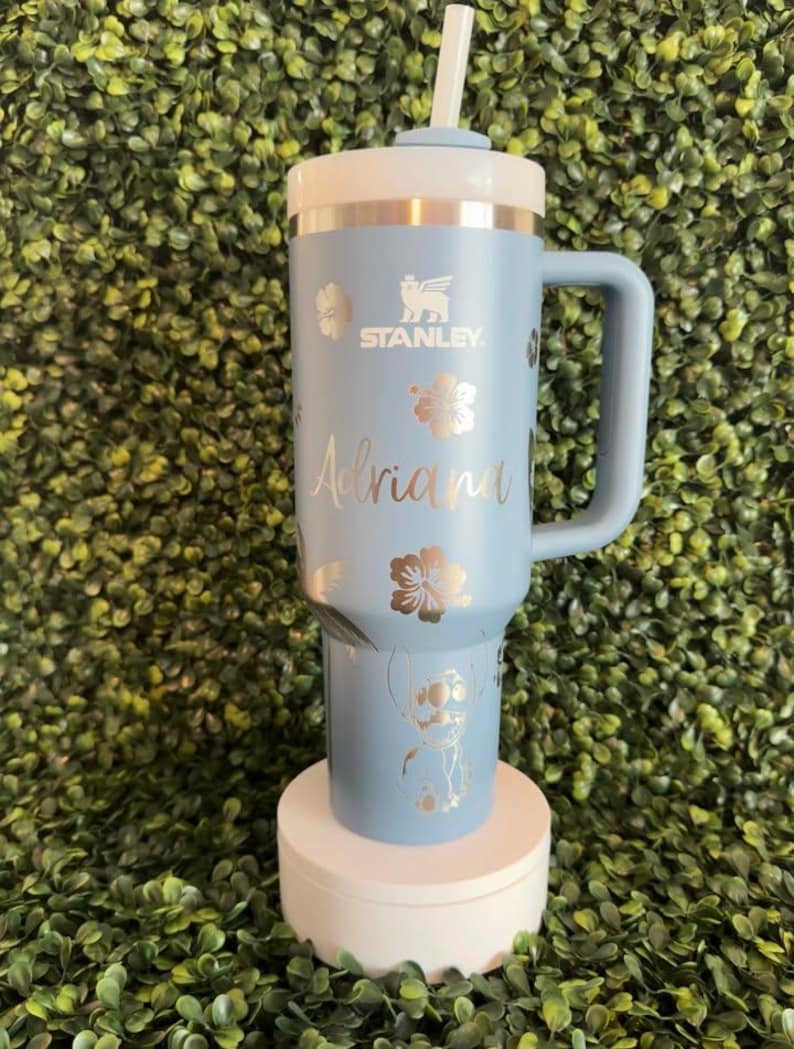 May include: A light blue Stanley tumbler with a white lid and handle. The tumbler features silver floral designs, the name "Adriana", and a cartoon character. It includes a white straw and is displayed on a white stand.