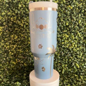 May include: A light blue tumbler featuring a gold-colored illustration of Stitch, the character from Lilo & Stitch. The tumbler includes the word "Stitch" and tropical designs. It has a white lid and a white straw.