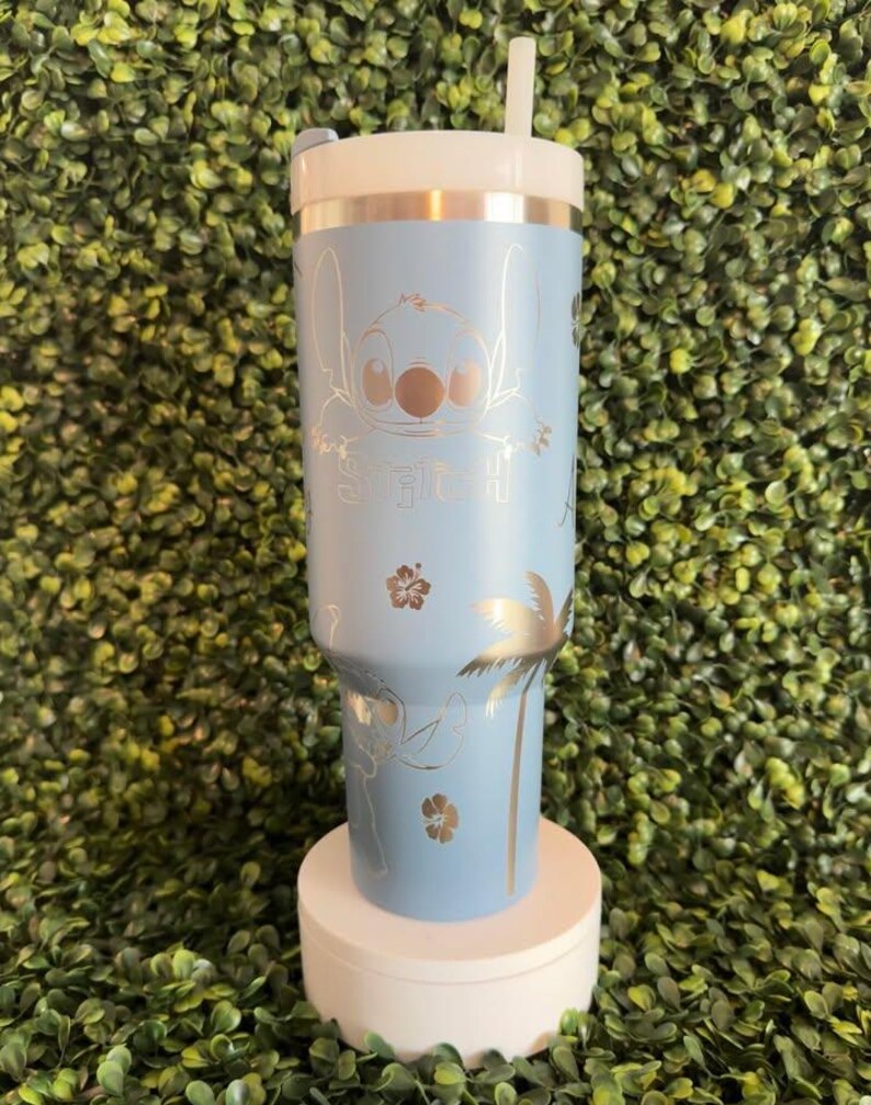 May include: A light blue tumbler featuring a gold-colored illustration of Stitch, the character from Lilo & Stitch. The tumbler includes the word "Stitch" and tropical designs. It has a white lid and a white straw.