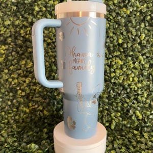 May include: A light blue insulated tumbler with a white lid and handle. The tumbler features a design with the words "Ohana means family", a sun, flowers, and a cartoon character playing a ukulele. Includes a white straw.