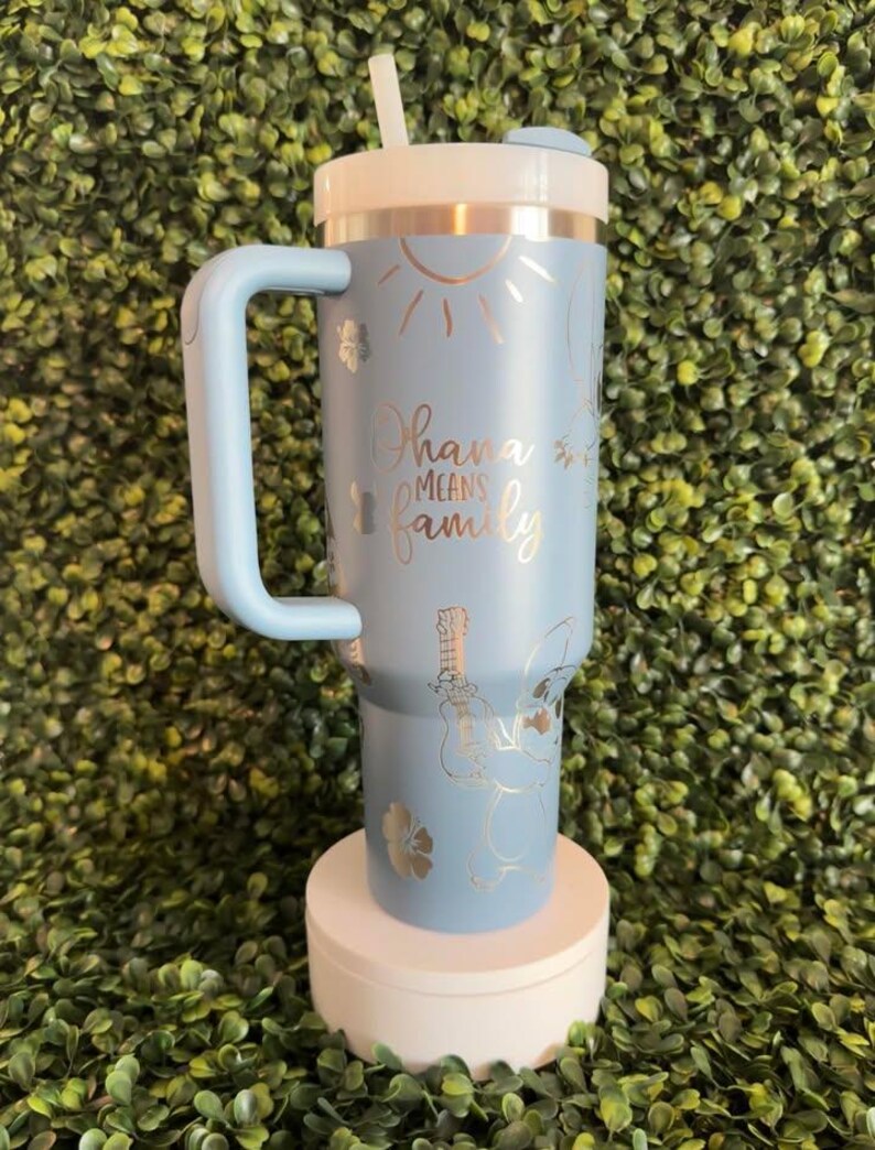 May include: A light blue insulated tumbler with a white lid and handle. The tumbler features a design with the words "Ohana means family", a sun, flowers, and a cartoon character playing a ukulele. Includes a white straw.