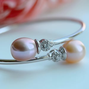 May include: A silver bypass bangle bracelet featuring two lustrous, light pink pearls at each end. Each pearl is accented by a small, circular cluster of clear, sparkling stones set in silver, creating a refined and balanced design on the open band.