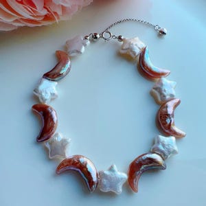 Op de afbeelding: Een delicate armband met afwisselende ster- en halvemaanvormige parels. De parels zijn wit en iriserend bruin, geregen op een zilveren ketting met een sluiting. Een uniek sieraad.