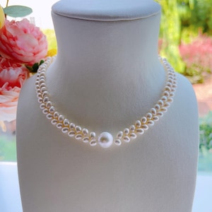May include: A delicate pearl necklace displayed on a white mannequin bust. The necklace features a double strand design with small pearls and a larger pearl pendant in the center. The background includes pink flowers and greenery.