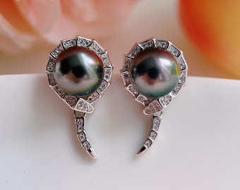 May include: A pair of silver-toned earrings featuring large, dark, iridescent pearls centered within a circular frame. The frame is encrusted with small, clear, sparkling stones and extends into a curved, tapered tail design.
