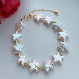 Op de afbeelding: Een delicate armband met stervormige parels met iriserende oppervlakken. De sterren zijn geregen met goudkleurige kralen en een delicate ketting, waardoor een hemels ontwerp ontstaat. De armband is verstelbaar.