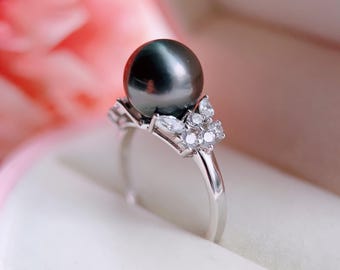 Könnte beinhalten: Ein silberfarbener Ring mit einer großen, runden, dunkelgrauen Perle in der Mitte. Das Band ist auf beiden Seiten mit einer Ansammlung kleiner, klarer, marquise- und rundgeschliffener Steine verziert, die ein symmetrisches Design um die Perle bilden.