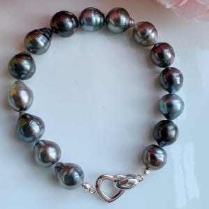 Op de afbeelding: Een donkergrijze parelarmband met een zilveren hartvormige sluiting. De parels hebben een onregelmatige vorm en een glanzende glans. De armband ligt op een witte ondergrond.