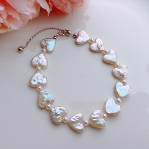 Op de afbeelding: Een delicate armband met hartvormige, iriserende parels. De armband is geregen met kleine, ronde parels tussen elk hart. De sluiting en ketting zijn zilverkleurig. De sieraden worden gepresenteerd op een wit oppervlak met een roze bloem op de achtergrond.