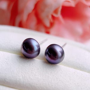 May include: A pair of dark purple pearl stud earrings. The pearls have a lustrous sheen and are set on silver posts. The background features a soft, blurred pink and white backdrop, highlighting the earrings.