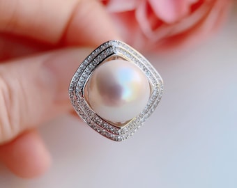 May include: A square-shaped ring featuring a large, lustrous pearl. The pearl is surrounded by a double halo of small, sparkling stones set in a silver-colored metal. The ring's design is elegant and eye-catching.