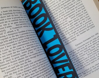 May include: A close-up of a book with a blue and black bookmark that says "BOOK LOVER". The book is open to a page with text about the journals of Lewis and Clark's expedition. The text describes their travels through the American West, including encounters with Native American tribes and the discovery of new plants and animals.