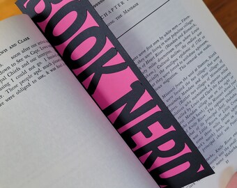 May include: A pink and black bookmark with the word "BOOK NERD" printed in bold white letters. The bookmark is partially inserted into a book with the text "CHAPTER V AMONG THE MANDANS" visible.