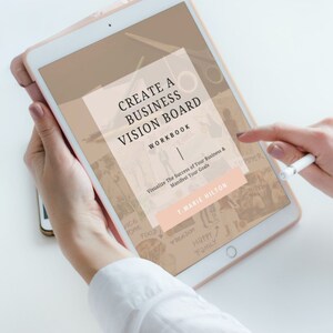 May include: A pink tablet computer displaying a digital workbook titled "Create a Business Vision Board Workbook" by T. Marie Hilton. The workbook is designed to help visualize the success of a business and manifest goals. The tablet is on a white surface.