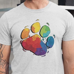 May include: A light gray t-shirt featuring a colorful paw print design. The paw print is filled with a rainbow of colors, including red, orange, yellow, green, and blue. The shirt is made of a soft material.