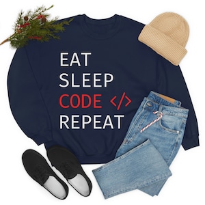 May include: Navy blue sweatshirt with white text that reads "Eat Sleep Code Repeat" with the word "Code" in red and angle brackets around it. The sweatshirt is laid flat on a white background with a pair of black trainers, a pair of blue jeans, and a beige beanie hat.