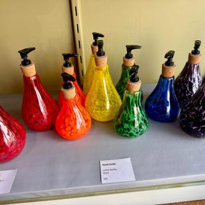 Puede incluir: Un surtido arcoíris de diez dispensadores de jabón de vidrio con tapones de corcho. Los dispensadores son de varios colores, incluyendo rojo, naranja, amarillo, verde, azul y morado. Los dispensadores están dispuestos en una fila sobre una superficie gris. El texto "David Smith Lotion Bottles Glass 4,00 €" es visible en una etiqueta blanca.