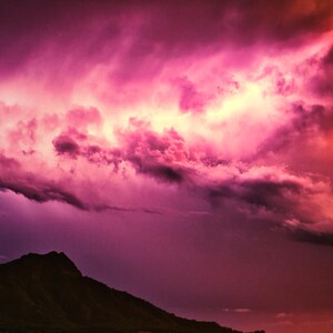 May include: Dramatic landscape featuring a vibrant sky dominated by shades of pink, purple, and orange. Dark, ominous clouds are illuminated by a flash of lightning. A silhouetted mountain range is visible in the foreground.