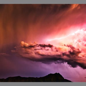 May include: A dramatic photograph of a thunderstorm with vibrant colors. The sky is filled with dark, swirling clouds, illuminated by flashes of lightning. The colors range from deep purples and oranges to bright pinks and reds, creating a striking visual contrast.