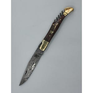 Peut inclure: Un couteau pliant doté d'une lame en acier damas au motif ondulé, d'un manche en bois sombre avec des mitres en laiton et d'un tire-bouchon en métal intégré. Le manche est incrusté d'un motif floral doré et d'une gravure en forme de lettre H.