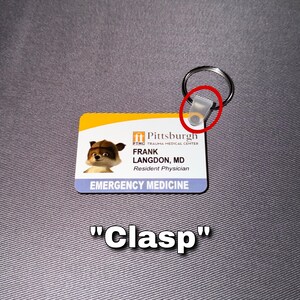 May include: A rectangular ID badge with a cartoon raccoon image, the text "FRANK LANGDON, MD, Resident Physician, EMERGENCY MEDICINE", and a Pittsburgh hospital logo. The badge is attached to a metal keyring with a clear plastic clasp.