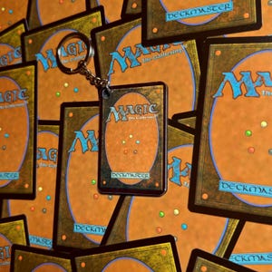 May include: A rectangular keychain featuring the back design of a Magic: The Gathering trading card, displaying the iconic orange and brown color scheme, the blue Magic logo, and the five-color mana symbol circle, attached to a silver metal ring.