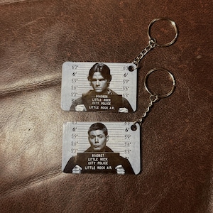 May include: Two metal keychains featuring mugshot-style images of men. Each keychain has a silver chain and ring. The mugshot background has height markings. The men hold signs that read "LITTLE ROCK CITY POLICE" and "LITTLE ROCK A.R."