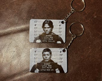 May include: Two metal keychains featuring mugshot-style images of men. Each keychain has a silver chain and ring. The mugshot background has height markings. The men hold signs that read "LITTLE ROCK CITY POLICE" and "LITTLE ROCK A.R."