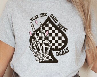 May include: Gray t-shirt with a black and white checkered spade design. A skeleton hand with pink lightning bolts is making a peace sign. The text "Play the hand you're dealt" is printed on the shirt.