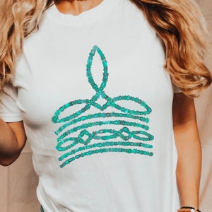 May include: White t-shirt featuring a teal-colored graphic design. The design appears to be an abstract crown or flame shape, composed of small, connected beads. The shirt is worn with a pearl necklace and bracelets.