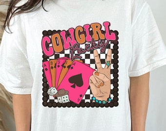 May include: White t-shirt with a graphic design featuring the text "COWGIRL Lucky" in pink and orange letters. The graphic also includes playing cards, dice, and a hand making a peace sign.