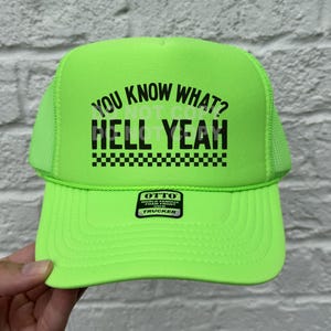 May include: A neon green trucker hat with black text that reads "You know what? Hell yeah" with a black and white checkered pattern underneath. The hat has a black label that reads "OTTO World Famous Foam Front Trucker".