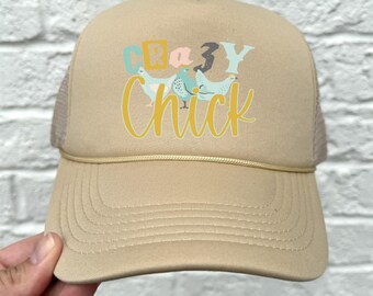 May include: A tan trucker hat with a mesh back. The front of the hat has a graphic of chickens and the text "Crazy Chick" in yellow.