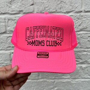 May include: Bright pink trucker hat with the text "CAFFEINATED MOMS CLUB" in black. The hat has a mesh back and a curved brim. The brand label "OTTO" is visible on the front.