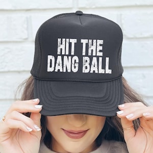 May include: Black trucker hat with the text "HIT THE DANG BALL" in distressed white lettering. The hat has a mesh back and a curved brim. A casual accessory for everyday wear.