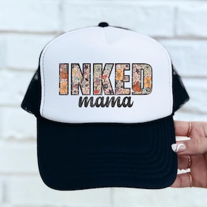 May include: A white and black trucker hat with a black mesh back. The front of the hat has the words "INKED mama" in black text with a colorful tattoo design behind the letters.