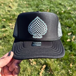 May include: Black trucker hat with a checkered ace of spades design outlined in turquoise. The hat has a mesh back.