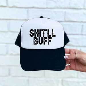 May include: A white and black trucker hat with a black mesh back. The front of the hat has the text "SHIT'LL BUFF" in black.