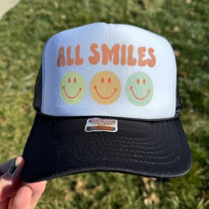May include: A white trucker hat with a black mesh back. The front of the hat has the words "ALL SMILES" in a retro font with three yellow smiley faces.