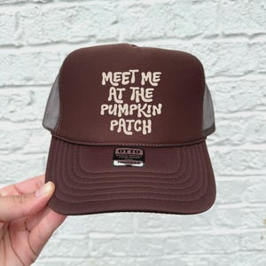 May include: A brown trucker hat with a mesh back and a curved brim. The front of the hat features the text "MEET ME AT THE PUMPKIN PATCH" in white, stylized lettering. The hat has a tag that reads "OTTO".
