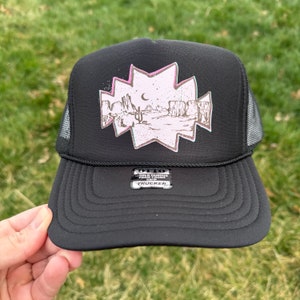 May include: Black trucker hat with a pink and green graphic of a desert landscape with cacti and mountains. The hat has a mesh back and a label that says "World Famous Foam Front Snapback Trucker".