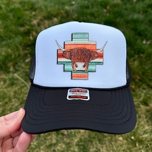 May include: A white and black trucker hat with a brown and white Highland cow head graphic on the front. The graphic is surrounded by a colorful striped pattern. The hat has a black mesh back and a black bill. The hat has a black label that says "WORLD FAMOUS FOAM FROM OTTO TRUCKER".
