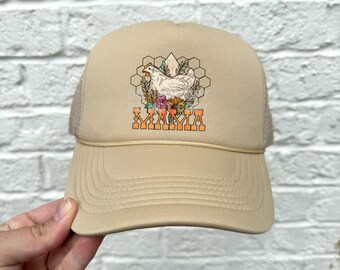 May include: A beige trucker hat with a white chicken and floral design. The hat has a mesh back and the words "Mama" are printed in orange on the front.