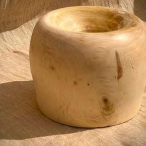 Puede incluir: Un cuenco de madera clara con una forma redonda y lisa y un patrón de veta de madera natural. El cuenco está vacío y tiene una ligera hendidura en el centro.