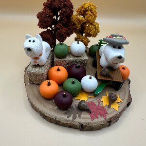 May include: A decorative fall-themed display featuring two white dog figurines, pumpkins in various colors, and miniature trees. The arrangement is set on a wooden slice base, with decorative leaves and hay bales.