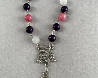 Op de afbeelding: Een kralenrozenkrans met afwisselend paarse, roze en witte kralen. Een zilverkleurige hanger toont een religieuze scène en een ovaal medaillon met een heiligenfiguur en het woord "ANDREW".