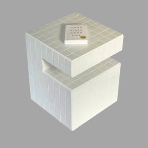 Op de afbeelding: Witte betegelde bijzettafel met twee planken. Een boek met de titel "Good Vibes, Good Life" ligt op de bovenste plank.