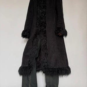 May include: A long black coat with faux fur trim on the collar, cuffs, and down the front. The coat is worn with dark wash denim jeans and black shoes. The coat appears to be made of a suede-like material.
