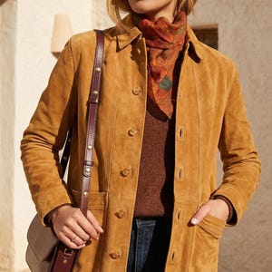 May include: A tan suede jacket with a button-up front, worn over a brown sweater and a patterned scarf. A brown leather crossbody bag with a long strap is also visible. The jacket has pockets and a classic collar.